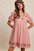 Woman wearing a pink dress with ruffled collar and embroidered hem on a beige background