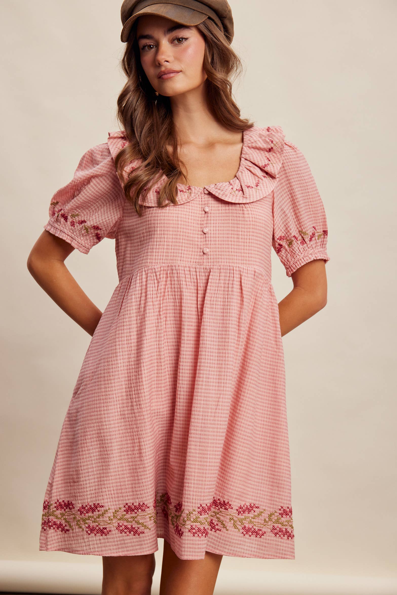 Woman wearing a pink checkered dress with ruffled collar and floral embroidery on a beige background